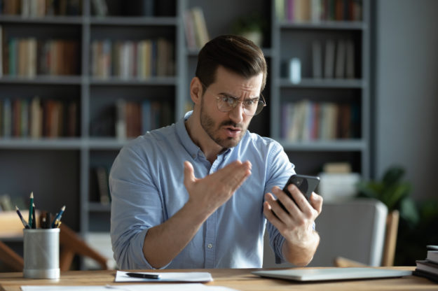 man sitting at desk, looking frustrated and gesturing towards smartphone