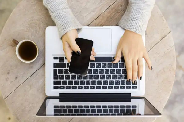 woman holding smartphone in right hand while typing on laptop with left hand