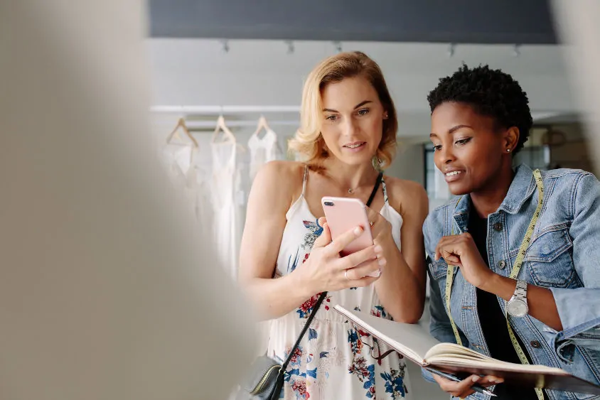 A female African-American tailor looks at a smartphone being held by a white patron inside clothing store