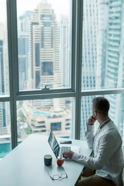 man staring out window at skyscrapers while working on laptop