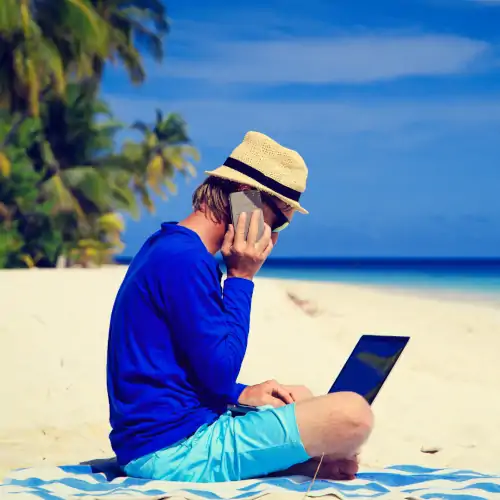 man sitting on towel at beach while talking on cell phone and typing on laptop