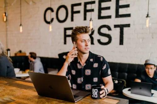 man on cell phone in coffee shop, sitting with laptop and holding mug with "80/20" written on it, with "COFFEE FIRST" written on wall in background