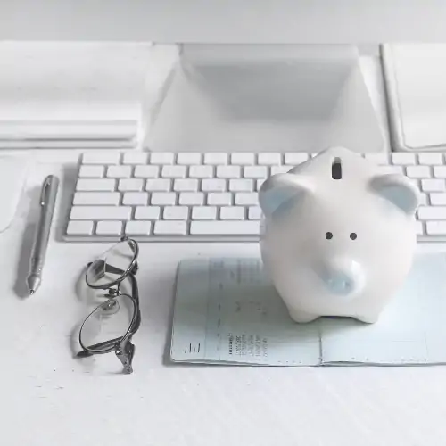 white piggy bank sitting on bills on desk with white keyboard, glasses, and pen