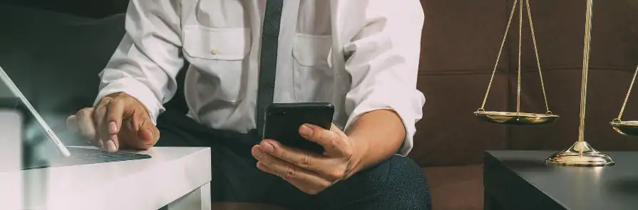 lawyer holding smartphone while working on laptop with scales on table next to them